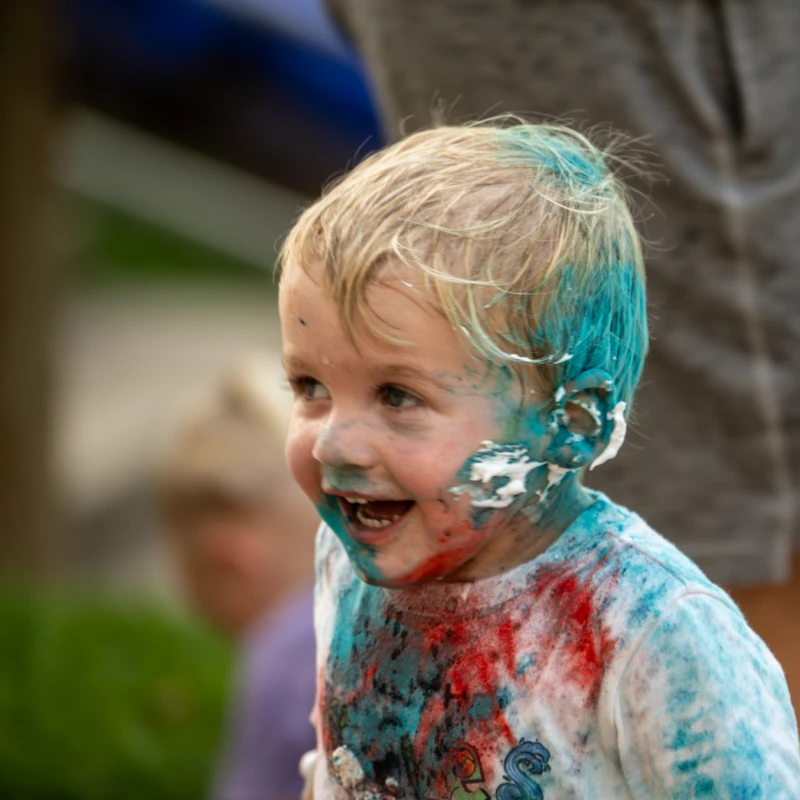 A happy young child with blonde hair laughs while covered in blue, red, and white frosting on their face, hair, and clothes.